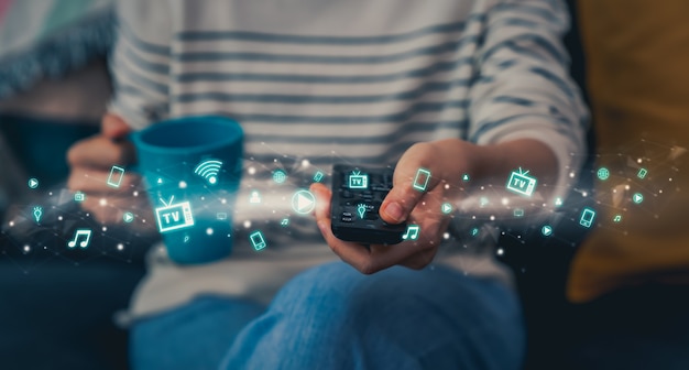 Woman sitting on the sofa and holding the TV remote to watch movies with multimedia icons on the screen on a relaxing day in the house.