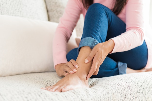 Woman sitting on sofa holds her foot injury, feeling pain.