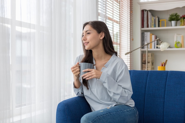 写真 ソファに座っている女性が窓際の景色を見て、日差しの中で最初の朝のコーヒーを楽しんでいます