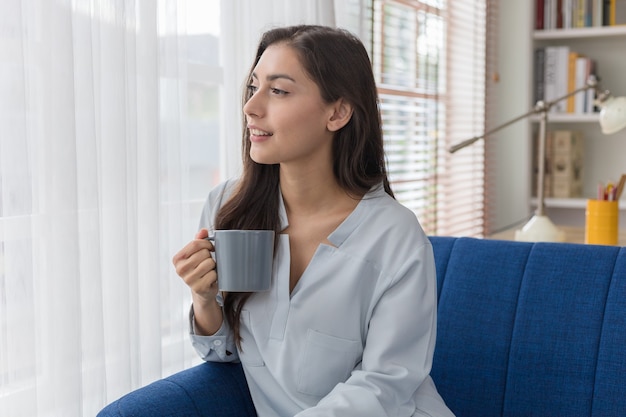 写真 ソファに座っている女性が窓際の景色を見て、日差しの中で最初の朝のコーヒーを楽しんでいます