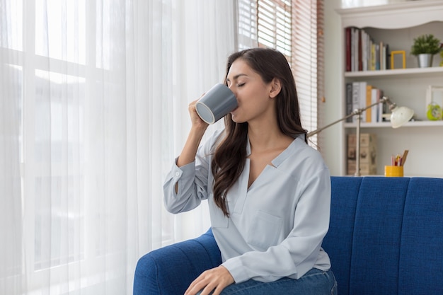 ソファに座っている女性が窓際の景色を見て、日差しの中で最初の朝のコーヒーを楽しんでいます