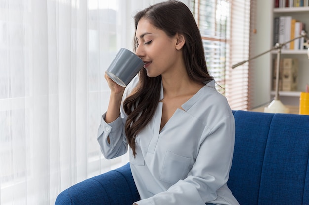 ソファに座っている女性が窓際の景色を見て、日差しの中で最初の朝のコーヒーを楽しんでいます