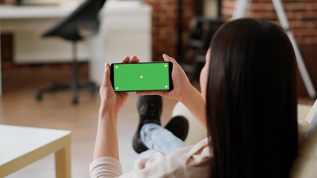 Woman sitting on sofa while holding smartphone with green screen mockup template background. Young adult person with modern touchscreen mobile phone having chroma key isolated display.