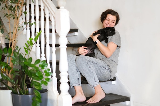 Woman sitting on stairs, using smartphone with black griffon or pug on her lap