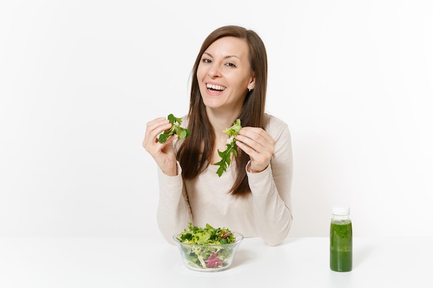 緑のデトックススムージー、ガラスのボウルにサラダ、白い背景で隔離の手でテーブルに座っている女性。適切な栄養、ベジタリアン料理、健康的なライフスタイル、ダイエットのコンセプト。コピースペースのある領域。