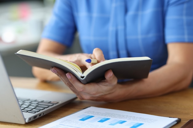 Woman sitting at table with laptop and documents and holding notebook for notes in her hands