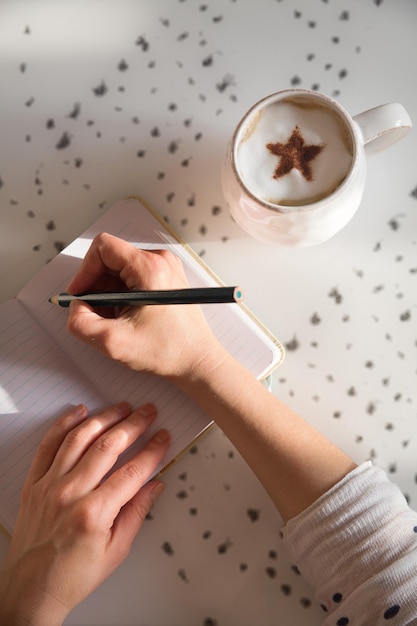 Woman sitting at the table writing in the notebook and drinking coffee in nice light home interior