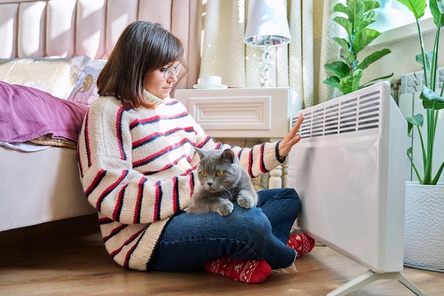 woman sitting with cat in a warm sweater warming near an electric heater