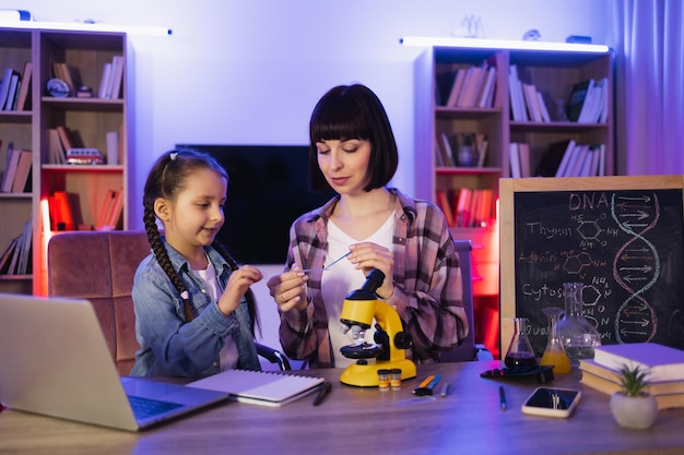 Woman sitting with girl at table and using microscope for studying sciences