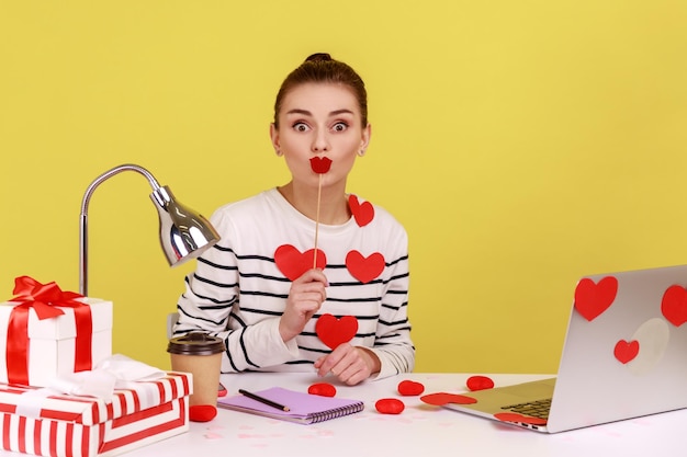 Woman sitting on workplace covered with heart sticks covering her mouth with paper lips