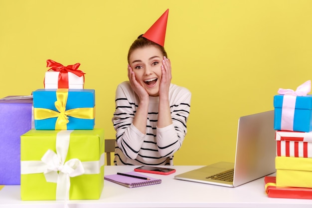 Woman sitting on workplace among present boxes wearing party cone celebrating her birthday at work