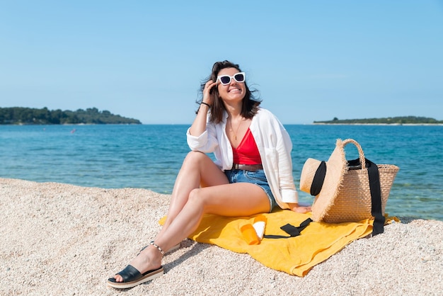 Женщина, сидящая на желтом одеяле на морском пляже в летнем наряде, защищает кожу от солнца в бутылках