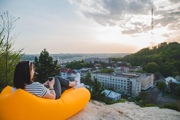 Woman sitting on yellow inflatable mattress drinking coffee enjoying sunset over city