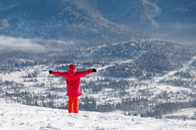 Woman in ski equipment takes in the sun\'s energy with her arms\
outstretched against the backdrop of blue mountain ranges. hooded\
jacket, red pants. healthy lifestyle. sports concept. selective\
focus.