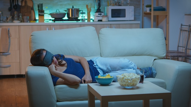 Donna che dorme con la maschera per dormire in soggiorno durante lo spettacolo televisivo. stanca esausta solitaria donna assonnata in pigiama che si addormenta sul comodo divano davanti alla televisione, chiudendo gli occhi mentre si guarda un film di notte.