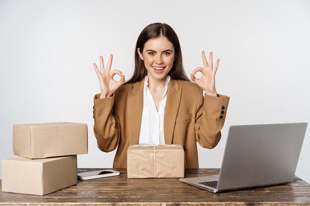 Donna titolare di una piccola impresa, seduta al tavolo con laptop, scatole di imballaggio, elaborazione degli ordini dei clienti, mostrando segno ok e ok e sorridente, in posa su sfondo bianco.