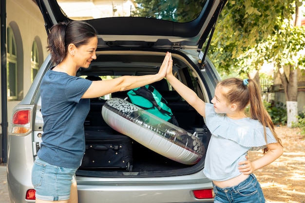 Woman and small girl doing highfive before leaving on holiday trip at seaside, loading baggage and inflatable in vehicle trunk. Mother and kid travelling by car during summer, journey adventure.