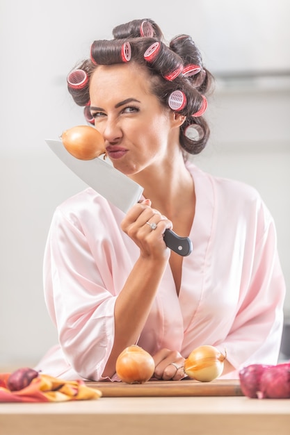 Woman smells onion and frowns while holding it stick to a knife in her hand.