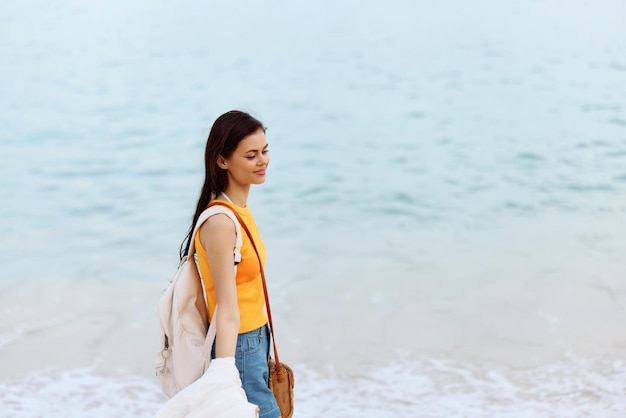 Woman smile with teeth after swimming in the ocean with a backpack in a wet yellow tank top and denim shorts walks along the beach summer vacation on an island by the ocean in Bali sunset
