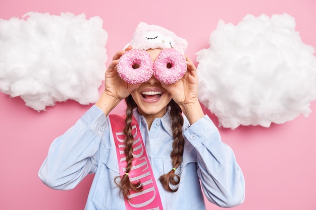 woman smiles gladfully cons eyes with glazed sweet doughnuts enjoys eating delicious bakery wears sleepmask shirt and birthday ribbon isolated on pink 