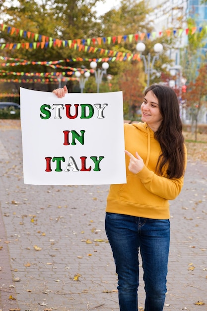 한 여성이 Study in Italy라는 문구가 적힌 포스터를 들고 미소를 짓고 있습니다.