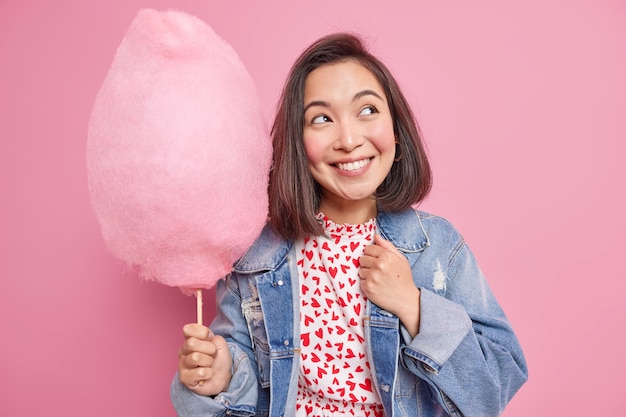 Photo woman smiles pleasantly looks away has dreamy expression dressed in denim jacket holds appetizing sweet candy floss wears stylish denim jacket