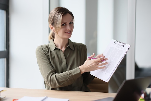 Женщина улыбается, показывает документ, подключенный к буферу обмена, и смотрит на концепцию камеры офиса