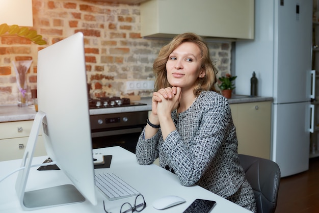 A woman smiles and works remotely on a desktop computer in her studio. A lady sits arms crossed during a video conference at home.