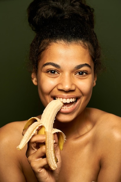 Foto donna che sorride alla telecamera, tiene e morde una banana gialla matura, posa isolata su green