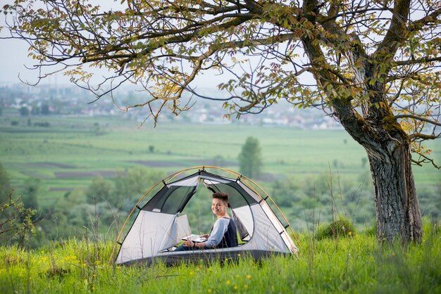 Woman smiling to the camera while using her laptop outdoors sitting in a tent on top of a hill under tree while camping