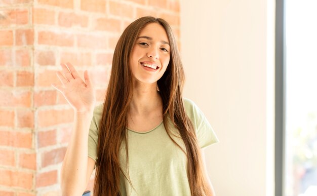 woman smiling happily and cheerfully, waving hand, welcoming and greeting you, or saying goodbye