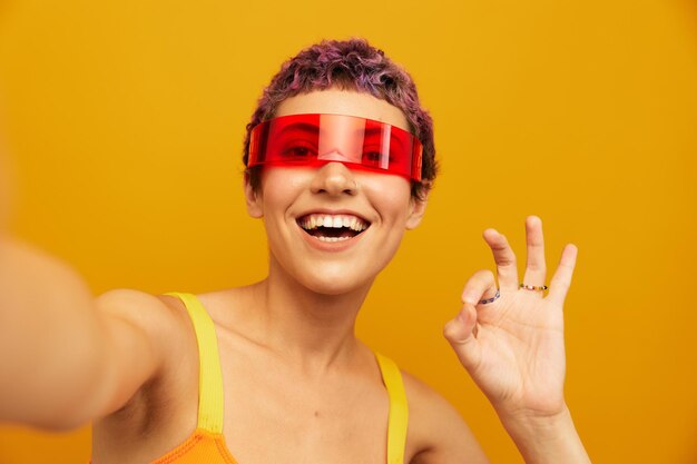 Woman smiling in unusual glasses millennial taking selfies in sportswear against an orange studio backdrop free space