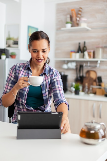 朝にタブレットpcを使用して笑って、熱い緑茶のカップを楽しんでいる女性