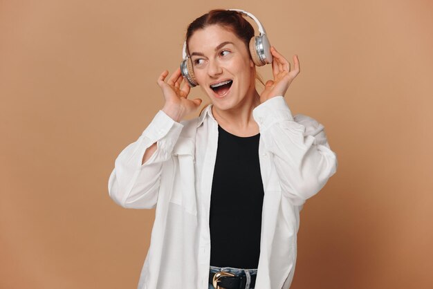 Woman smiling with braces on her teeth and listening to music in headphones on a beige background