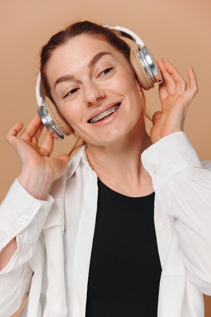 Woman smiling with braces on her teeth and listening to music in headphones on a beige background