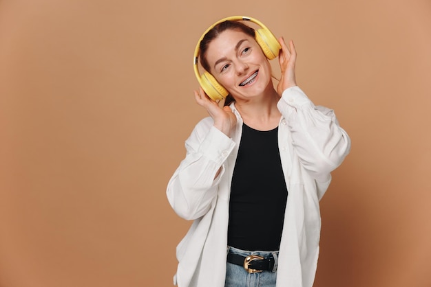 베이지색 배경 에 헤드폰 을 착용 하여 음악 을 듣고 있는 여성 이 치아 에 브레이크 를 착용 하여 미소 짓고 있다