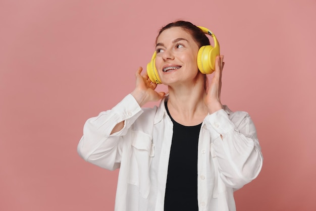 Woman smiling with braces on her teeth and listening to music in headphones on pink background