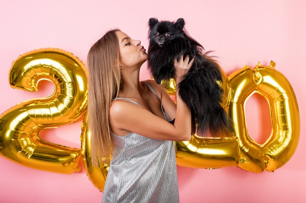 Woman smiling with japanese spitz black dog and golden 2020 new year balloons isolated over pink