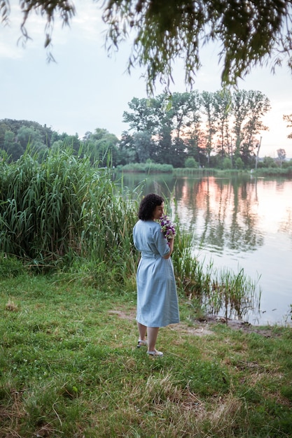 Woman sniffs flowers by lake at sunset