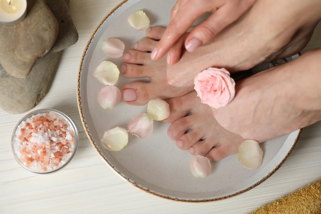 Woman soaking her feet in bowl with water flower and petals on white wooden floor top view Pedicure procedure