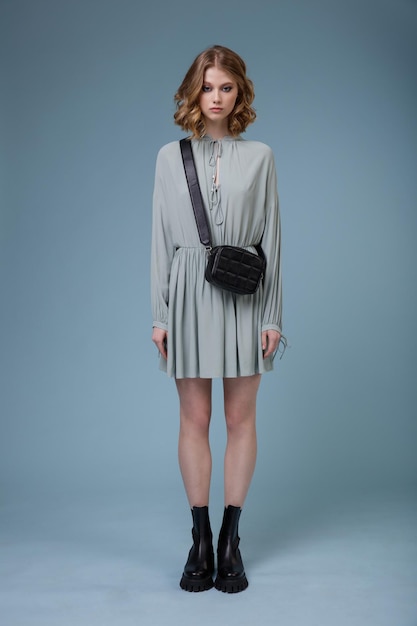 Woman in soft blue gray short dress boots black handbag posing on blue background