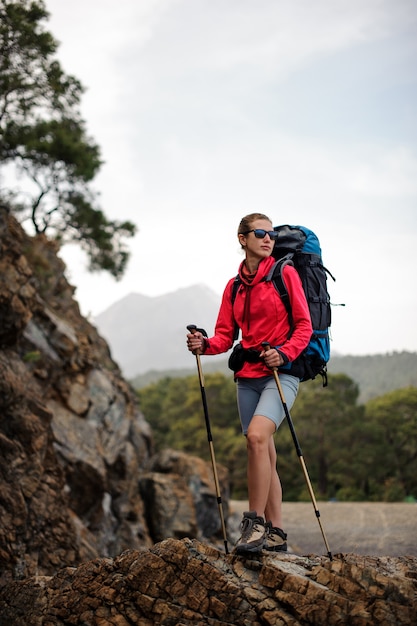 Woman in special wear standing with hiking backpack and sticks on the rock