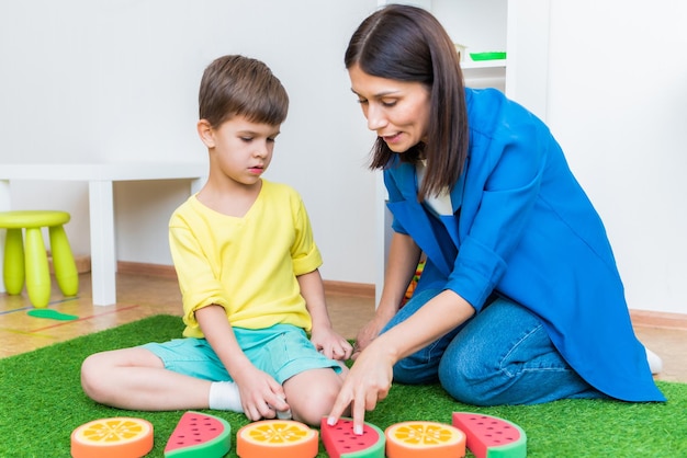 A woman speech therapist deals with a boy and performs an exercise to correct the speech apparatus by playing on the floor