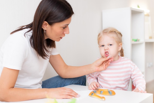 Woman speech therapist helps cute girl to learn correct pronunciation and competent speech in her office