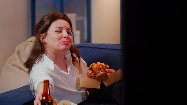 Фото Женщина проводит свободное время с едой на вынос и телевизором, сидя в гостиной. кавказский взрослый ест гамбургер и пьет пиво, смотрит телевизор после работы. человек, наслаждающийся едой