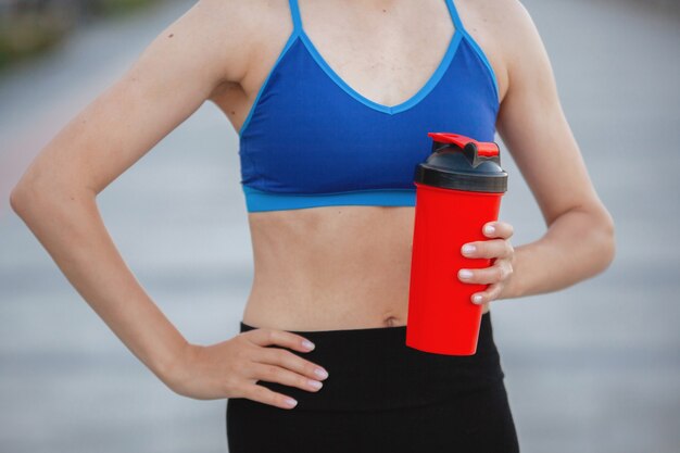 Foto donna in un top sportivo con un bicchiere di nutrizione sportiva durante una sessione di allenamento in una strada cittadina in estate. avvicinamento.