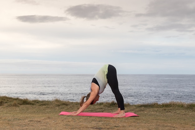 Woman in sportswear doing yoga outdoor on a pink mat with the sea