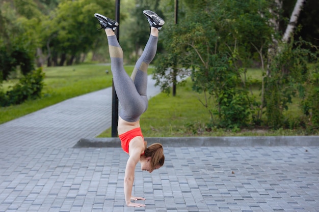 Woman in sportswear is standing on her hands in a gym in a summer city park.