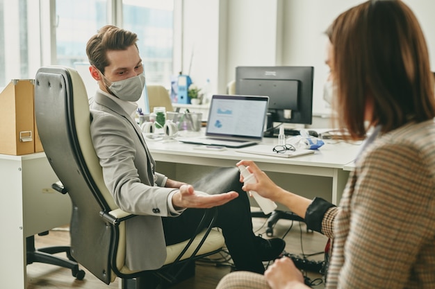 Woman spraying sanitizer onto colleagues hands while they working in half-empty office