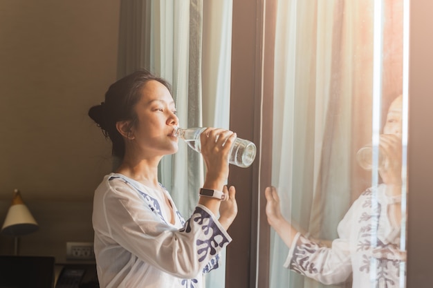 Woman stand next to window drinking water from bottle in nature light.